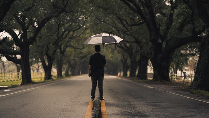 A man standing in the middle of the street, facing away and holding an umbrella